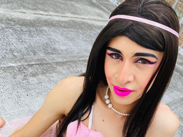 Catalina Castro, com batom rosa, sobrancelhas pretas grossas, delineador rosa, colar de pérolas, tiara rosa, cabelo preto longo, vestido rosa, sentada em uma superfície prateada texturizada, olhando para a câmera..