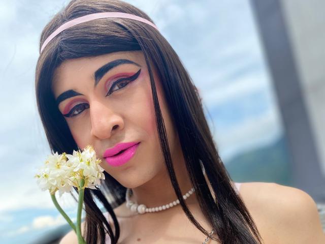 CatalinaCastro has pink lipstick, white flowers, and a pearl necklace, with long brown hair, pink headband, and bold makeup, holding a flower near her face.