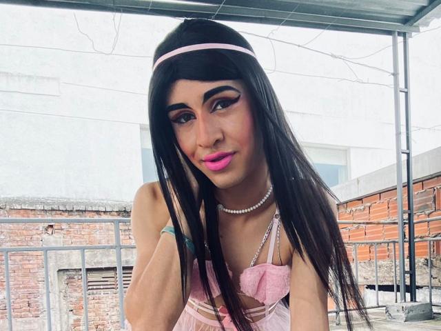 Catalina Castro mit langem, schwarzem Haar, einem rosa Haarreif, pinkem Lippenstift, einer Perlenkette, einem pinken Kleid und pinkem Lidschatten, die nach vorne gebeugt ist, kleine Brüste zeigt, niedlich aussieht, auf einem Dach..