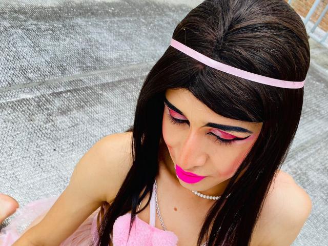 Catalina Castro usa batom rosa, maquiagem marcante nos olhos e uma tiara rosa. Ela está vestindo um vestido rosa e fofo, e um colar de pérolas. Seu cabelo escuro está solto, e ela está olhando para baixo..