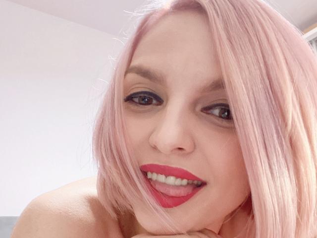 DouceAmelia with pink hair, red lipstick, and a playful smile, looking at the camera with her tongue slightly out. Her nose is pierced. She has black eyeliner.