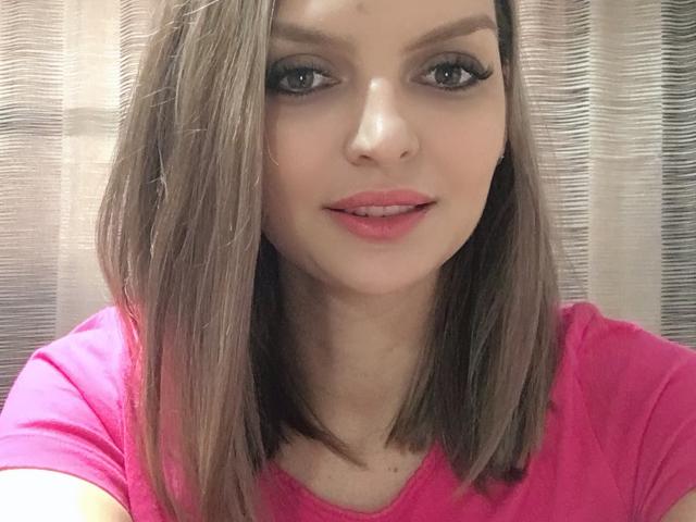 DouceAmelia has straight brown hair, pink lipstick, and blue eyes, wearing a pink shirt. She's smiling with a slight nose and visible teeth. Her tits are small and cute.