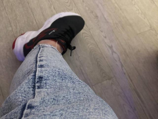 LadyMiller's ass in faded blue jeans, wearing black Nike sneakers with red accents, standing on wooden floor. Sneaker's white sole and black laces are visible.