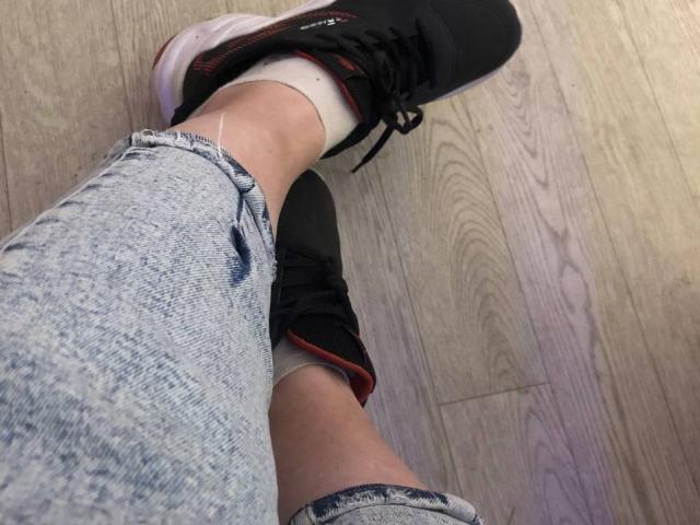 LadyMiller's legs crossed at the ankle, wearing black and pink sneakers with red trim, and light blue, slightly frayed jeans, on a wooden floor.