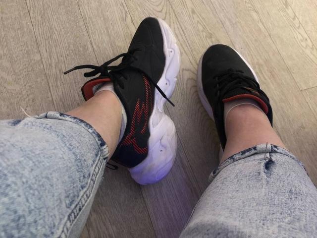 LadyMiller's legs in faded denim jeans, wearing black and white sneakers with red accents, standing on wooden floor. Her ankles are slightly exposed.