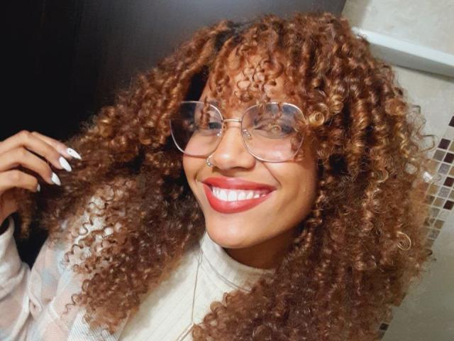 Cinthia Walker, con cabello castaño rizado, usando gafas, lápiz labial rojo y un cuello alto blanco, sonriendo con la mano en el cabello, mostrando sus uñas blancas..