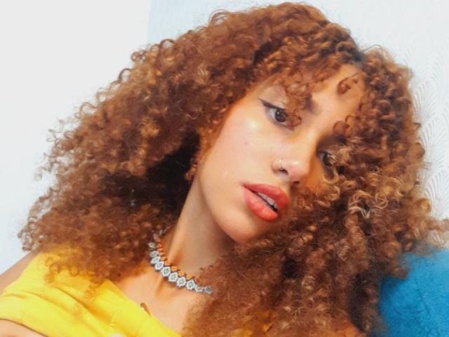 CinthiaWalker with curly brown hair, yellow top, and sparkly necklace, has full lips with red lipstick, and a nose piercing. She's looking to the side with a slight pout.