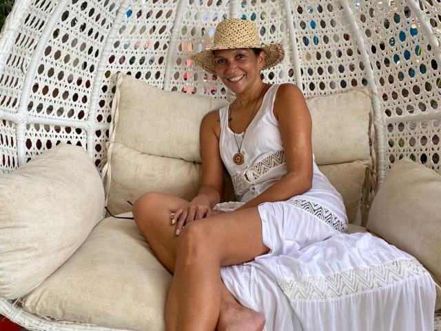 MatureJennyX with tan skin, smiling, wearing a white dress, straw hat, and necklace, sitting on a white wicker chair with beige cushions, legs crossed, displaying her ass.