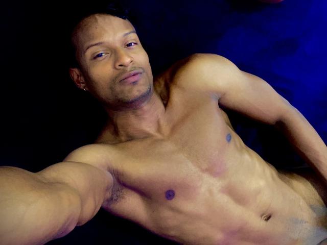 JacksonCooper's nude selfie shows a muscular, tan-skinned guy with a shaved head, small nipples, and a relaxed, confident expression, taking a selfie against a dark blue background.