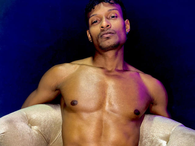 JacksonCooper, shirtless with a muscular, shiny brown chest, small dark nipples, and a slight smirk. Sitting on a beige, tufted chair with purple and blue lighting.