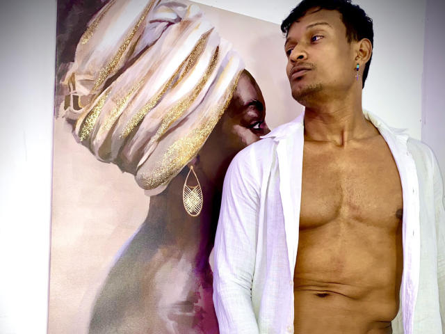 JacksonCooper, shirtless with a white button-up, stands next to a painting of a woman with glittery gold headwrap and earrings, showing her side profile, looking away.