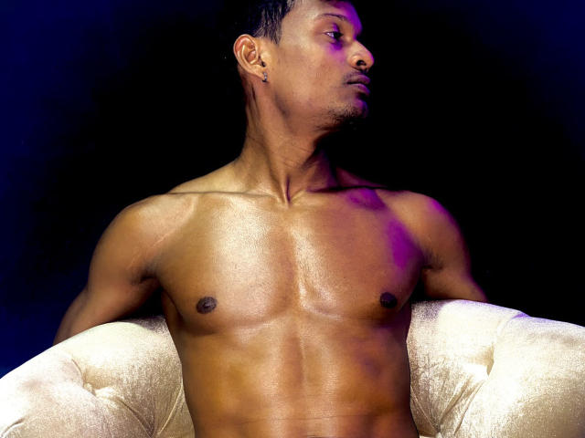 JacksonCooper's shiny, muscular, dark brown chest and abs are prominent, sitting on a white, tufted chair with purple lighting. He has short black hair and small black earrings.