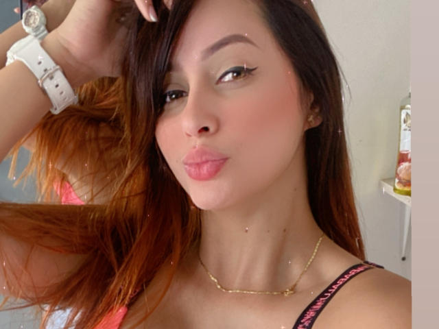 EmilyParkerr with pouty pink lips, brown eyes, and long brown hair, wearing a pink and black strap top, white watch, and gold necklace, looking seductive.