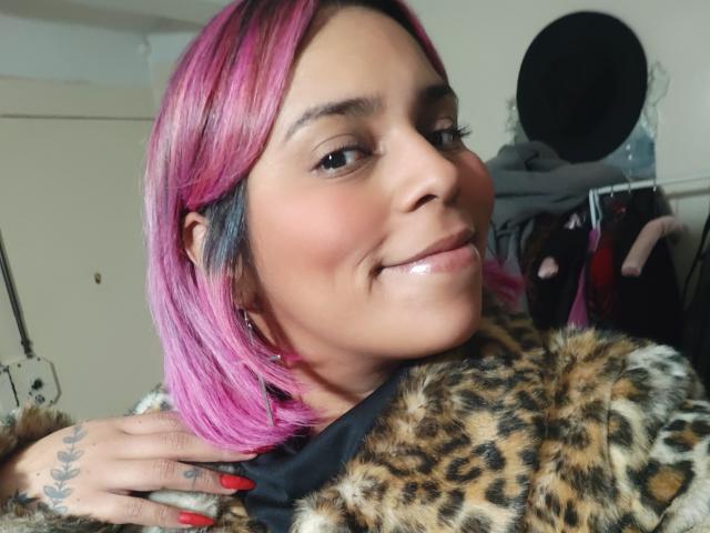 Pimentão com cabelo rosa sorrindo, facesitting em pele de impressão de leopardo, mão com unhas vermelhas e tatuagens sobre o ombro.