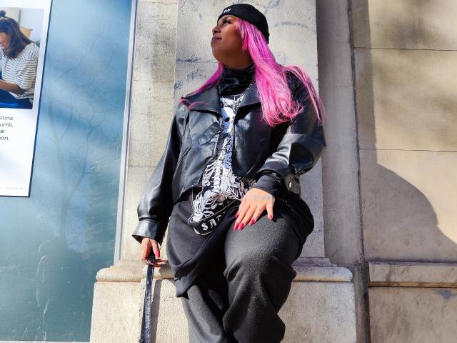 Chili with bright pink hair, wearing a black hat and oversized black outfit, leaning against a building, looking upwards.