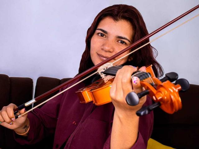 BulmaHot with long dark brown hair, holding a violin, has a small tattoo on her right hand, wearing a maroon shirt, and smiling softly.