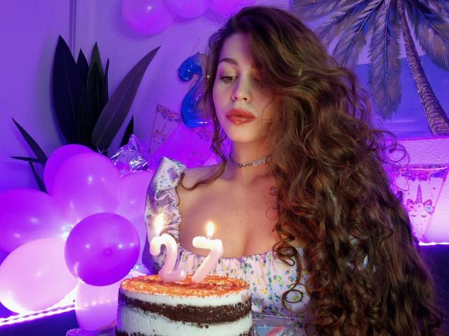 ValeriMair with long curly brown hair, wearing a choker, and a floral dress, holding a cake with lit candles, surrounded by purple balloons, in a purple-lit room.