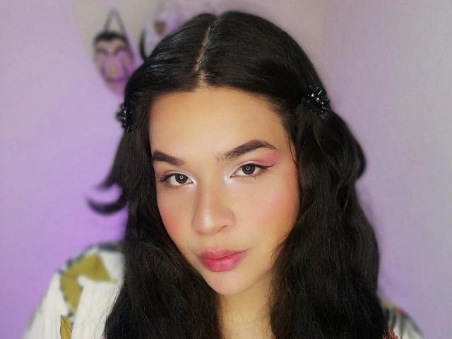 Antonela, mit langen schwarzen Haaren, rosa Lidschatten und rosa Lippenstift, trägt ein Oberteil mit Blumenmuster und hat einen niedlichen, sanften Gesichtsausdruck sowie einen leichten Rosaton auf ihrer hellen Haut..