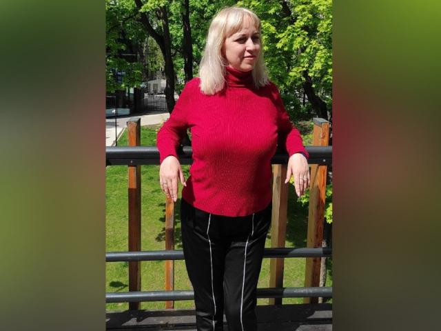 AnaBerry, eine blonde Frau, trägt einen roten Rollkragenpullover, schwarze Hosen und steht auf einem hölzernen Balkon. Ihre Arme ruhen auf der Reling. Im Hintergrund sind grüne Bäume zu sehen. Es ist sonnig und sie hat einen ruhigen Gesichtsausdruck. Sie hat kleine Brüste und eine mittelgroße Statur. Ihr Outfit ist leger..