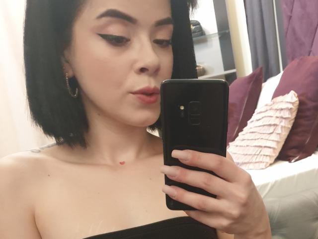 EvaLaurence con un caschetto nero, eyeliner allungato, orecchini a cerchio, che tiene in mano il telefono, sta scattando un selfie allo specchio, labbra rosate, un piccolo tatuaggio a forma di cuore rosso sulla clavicola, top nero senza spalline, cuscini viola sullo sfondo..