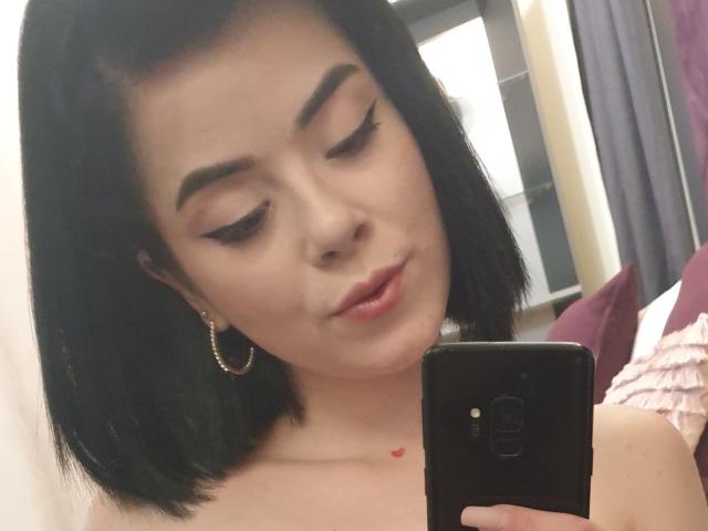 EvaLaurence, con un corte de pelo bob negro, aros grandes, y delineador de ojos, sosteniendo un teléfono, mirando hacia abajo, con una ligera sonrisa, un pequeño tatuaje de corazón rojo debajo de la clavícula, piel pálida, maquillaje sutil, busto medio..