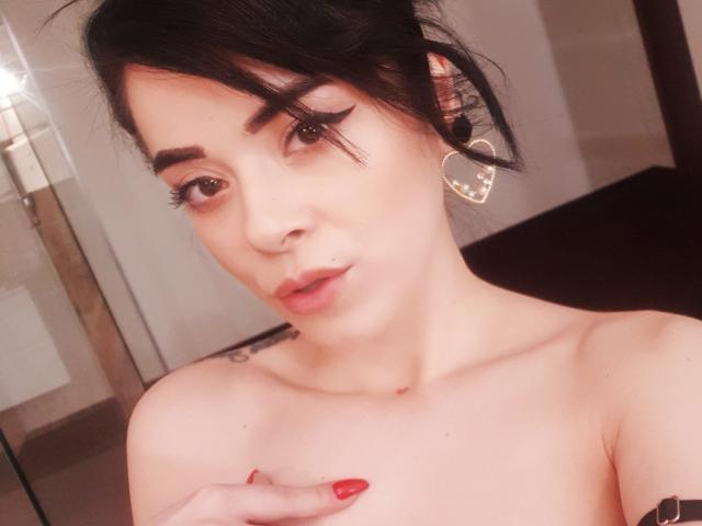 EvaLaurence has dark hair, thick eyebrows, and heart-shaped earrings. She's topless with red nails, pouty lips, and a tattoo on her right shoulder. Her nipples are visible.