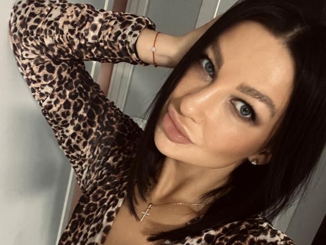 KristenStark with blue eyes, wearing a leopard print top, has her black hair down, looking at the camera with a slight smile, hand in her hair, small earrings.