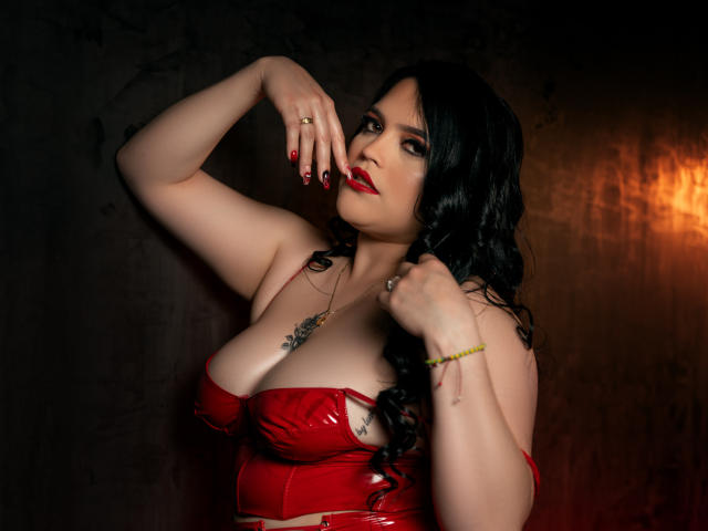 Maeve Borch con una maglia di lattice rossa, che mette in mostra un seno generoso, capelli neri ondulati, rossetto rosso, smalto per unghie rosso, una mano sul petto, un braccialetto giallo, un anello e un tatuaggio, con un'aria seducente..