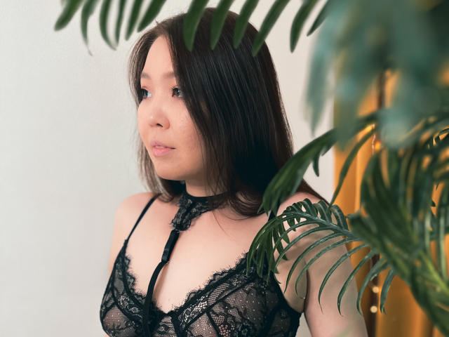NatzukoShi with medium tits, wearing black lace lingerie, has fair skin, brown hair, and a subtle smile, framed by green plant leaves. She's looking to the side.