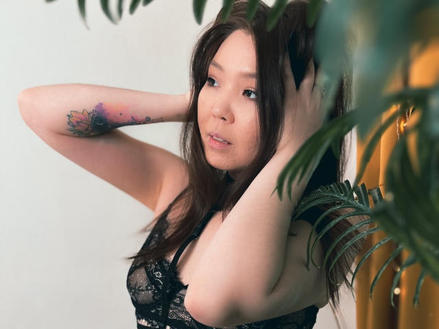 NatzukoShi with long brown hair, looking at camera, hands in hair, wearing black lace bra, tattoo on left arm, green leaves in foreground, soft smile, light skin.