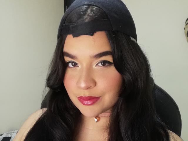 AntonelaPark with dark, wavy hair, wearing a black baseball cap, has pink lipstick, black eyeliner, and a small orange necklace, smiling slightly at the camera.
