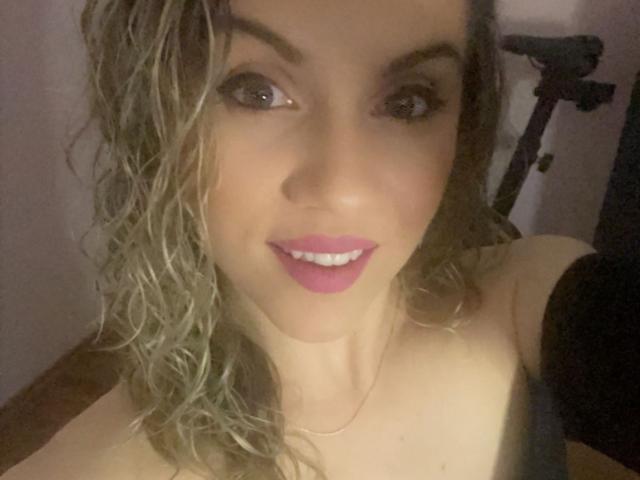LucilleXSugar a les cheveux blonds bouclés, du rouge à lèvres rose, et elle sourit, ses seins étant légèrement visibles. Elle a les yeux marrons et un teint clair, et elle porte un soutien-gorge à bretelles noires..