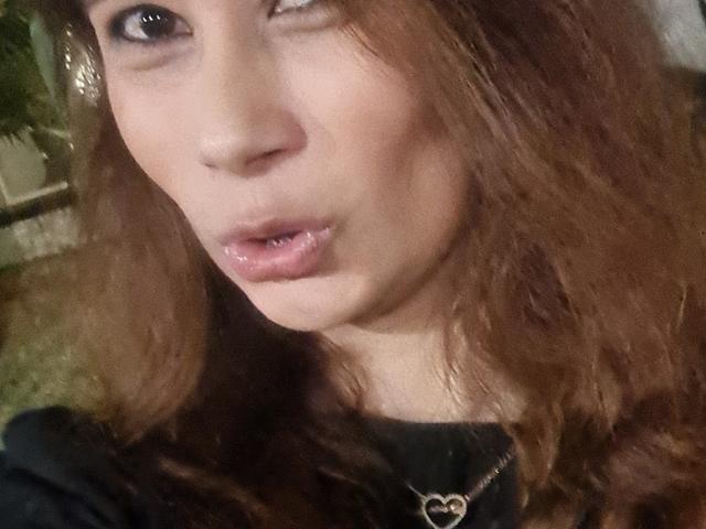 GiovannaSweet tem cabelos castanhos ondulados, pele clara e um lábio inferior com piercing. Seus lábios rosados estão ligeiramente entreabertos. Ela está vestindo uma camisa preta com um colar de coração..