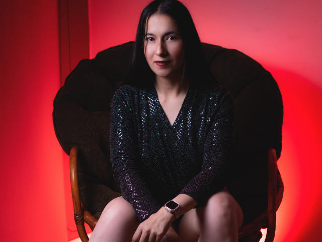 AnneStone with black sequined dress, sitting on brown chair, red background, black straight hair, red lipstick, smartwatch on left wrist, slightly smiling, legs together, arms resting on knees.