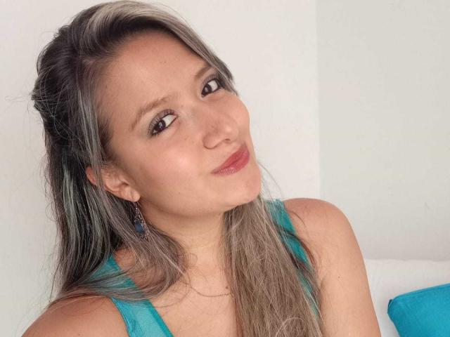 ValeriaHunter69 has light brown skin, long grayish-brown hair, and is wearing a blue tank top with matching earrings. She's smiling slightly and looking to the side.