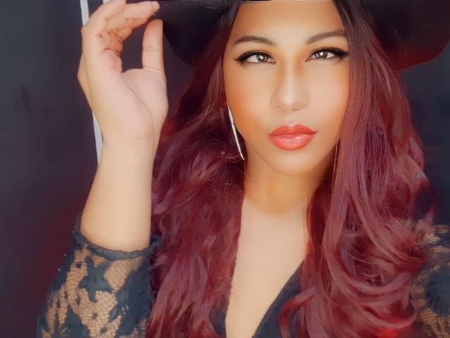 SakuraRiver, con i capelli rossi ondulati, una maglia nera in pizzo e rossetto rosso, tiene in mano un cappello nero, indossa orecchini a cerchio argentati, guarda direttamente la fotocamera con un'espressione sensuale..