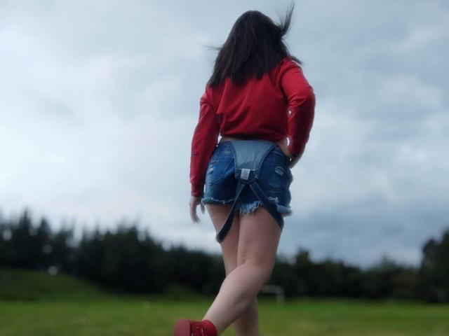 Kkatalina's ass is in blue denim shorts, she's wearing a red top, red socks, and her black hair is blowing. She's standing in a grassy field with trees.