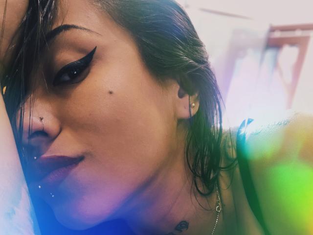 LunaCandy con eyeliner scuro, piercing al naso e orecchini, capelli castani leggermente umidi, un sorriso appena accennato, un tatuaggio visibile e una maglia nera, su uno sfondo colorato e sfocato..