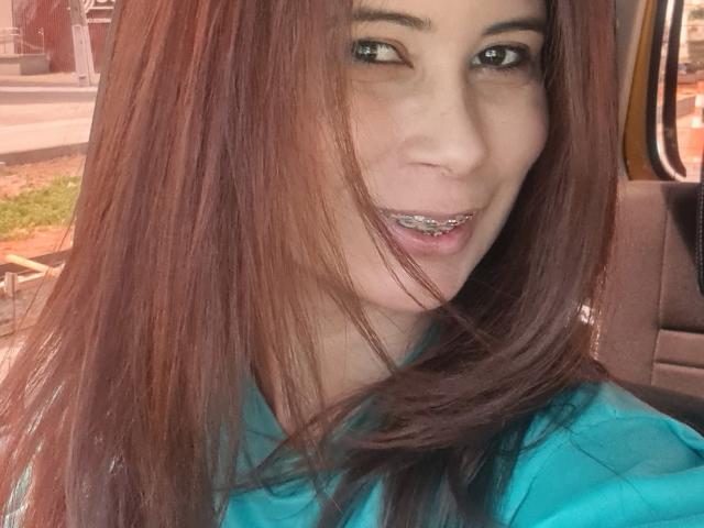 GiovannaSweet has brown hair, silver grill, and green eyes, wearing a teal shirt, with a slight smile in a car. Her ass is not visible.