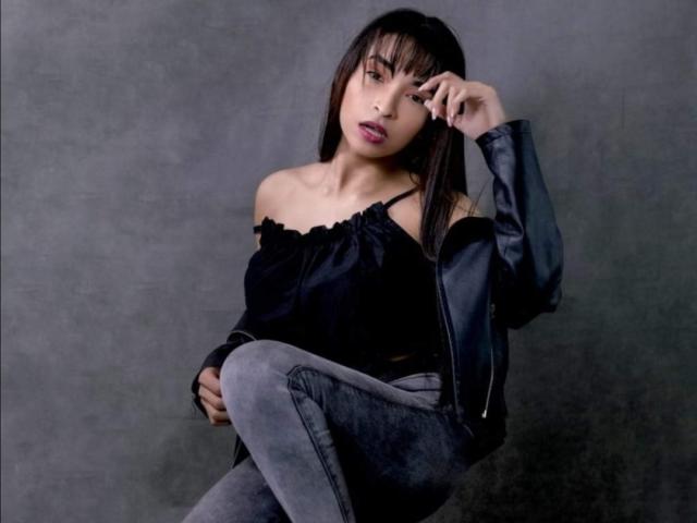 SarahyJhones, con senos pequeños, vestida con una blusa negra con un hombro descubierto, jeans grises y una chaqueta de cuero negra, sentada con una mano tocándose la frente, luciendo ligeramente aburrida..