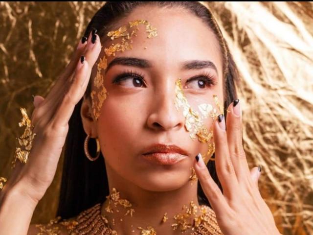 O rosto de SarahyJhones está coberto de folha de ouro, as mãos estão na cabeça, olhando para cima com olhos escuros, usando brincos de argola e com batom nude brilhante. Suas unhas são pretas com pontas douradas..