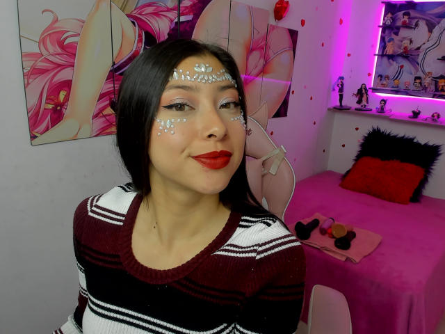 SofiLenox, con lunghi capelli neri, rossetto rosso e brillantini sul viso, indossa un maglione a righe marrone e bianco, sorridendo in una stanza illuminata di rosa, con poster di anime alle pareti..