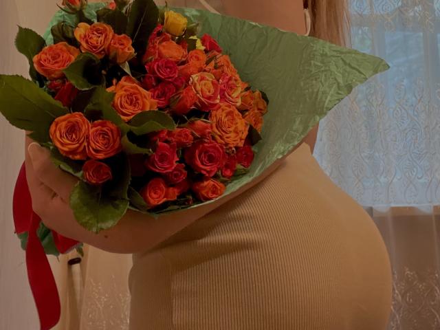 SugarrX tiene in mano un bouquet di rose arancioni e rosse, indossa un abito beige a coste, con il sedere leggermente visibile, la carta da imballaggio dei fiori è verde e c'è un nastro rosso..
