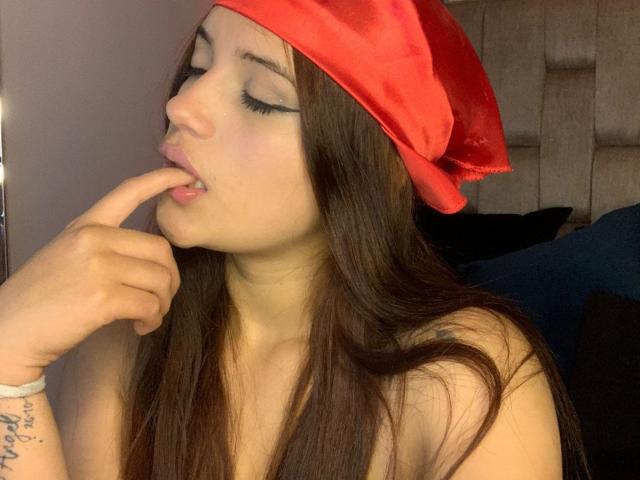 HelenTay, com seus longos cabelos castanhos, usando um chapéu de Papai Noel vermelho de veludo, tem o dedo na boca, exibindo seu delineador preto e marcante, e uma pulseira branca..