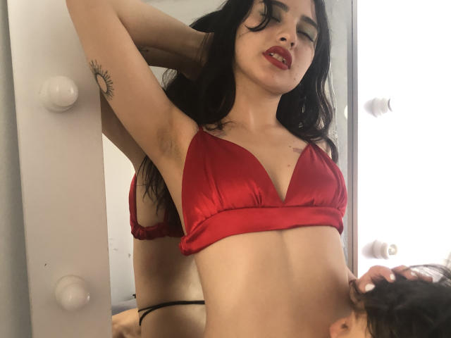 DamianMaya, com cabelo preto, sutiã vermelho e tanga preta, com as axilas visíveis, braços levantados, cabeça inclinada, lábios entreabertos, tatuagem no braço esquerdo, reflexo no espelho, cabeça de homem no canto inferior direito..