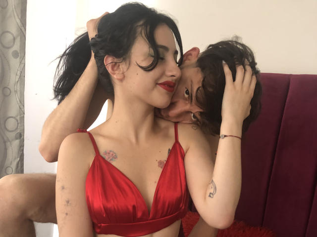 DamianMaya, com cabelo preto, sutiã de cetim vermelho, tatuagens e batom vermelho, sentada em uma cadeira magenta, enquanto outra pessoa com cabelo castanho encaracolado beija seu pescoço..
