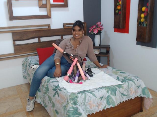 CataleyaX69, brown-skinned woman with black hair, sitting on bed with floral sheets, wearing jeans and tan top, smiling, surrounded by pink and black dildos, white towel beneath.