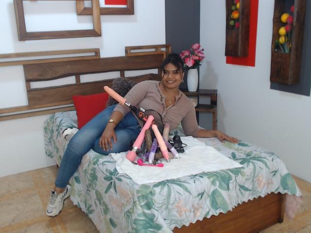 CataleyaX69, dark-skinned woman, laying on bed with floral sheets, wearing pink shirt, blue jeans, white sneakers, holding pink and purple dildo toys, smiling.