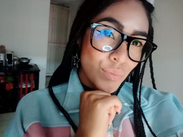 StellaBeckker with dark braids, black glasses, and light blue shirt, puckering lips, hand near chin, reflection in glasses showing blue eye, slight smile, pale skin, casual selfie.