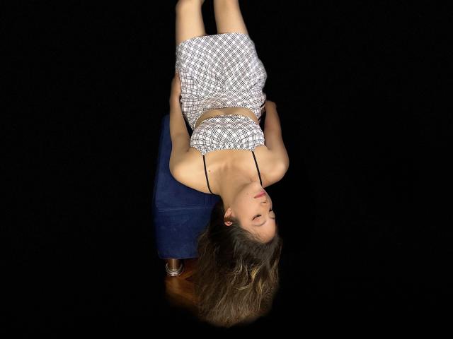 EvelinHotty, de cabeça para baixo, com cabelo castanho ondulado, seios pequenos e um vestido xadrez, pendurada em uma cadeira azul, contra um fundo preto, mostrando uma expressão relaxada e fofa..